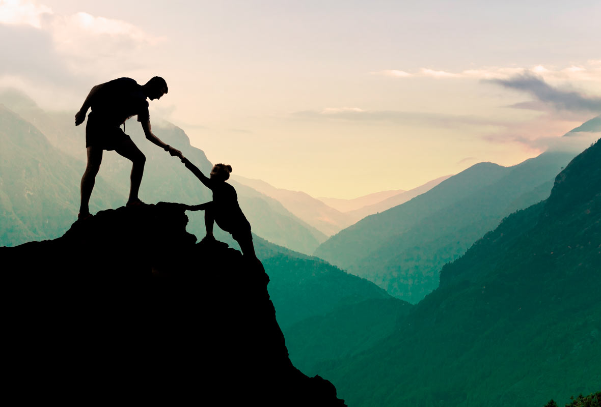 two people climbing a mountain top. The one at the top lends a hand to the other to make the final climb. 