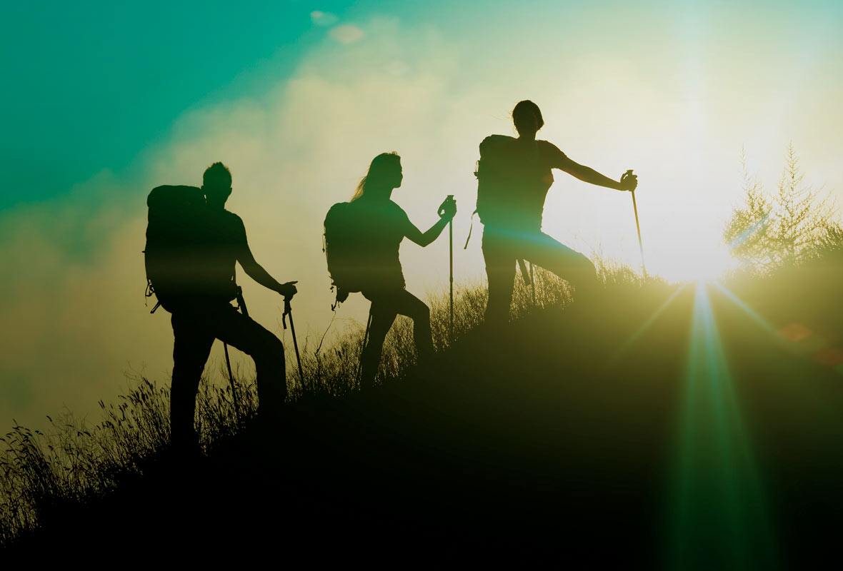 Three people hiking with walking-poles up a hill with the sunlight beaming in the background